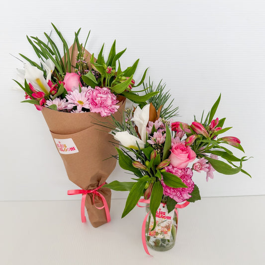 Two floral bouquets, one wrapped in brown kraft paper with a pink ribbon, and the other is arranged in a clear glass jar with a pink ribbon.