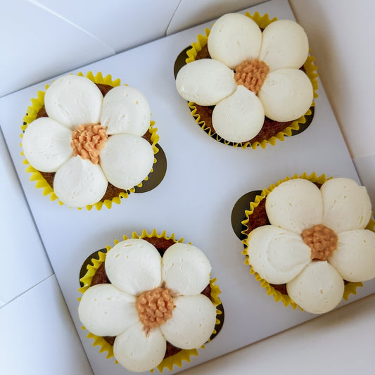 Four carrot cupcakes with white frosting petals inside a white box.