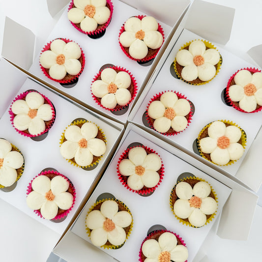 Four white bakery boxes arranged in a grid, each containing four decorated cupcakes topped with large cream-colored flower-shaped frosting pieces and small orange centers.