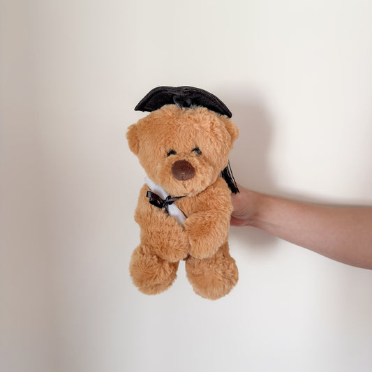 A plush brown teddy bear wearing a black graduation cap and holding a white diploma.
