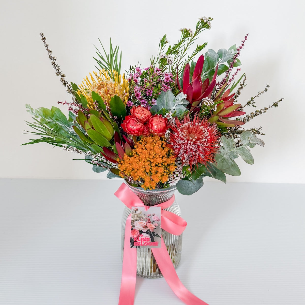Colourful bouquet of native flowers in a glass ribbed posy vase with a pink ribbon on a white background, available for delivery around Perth